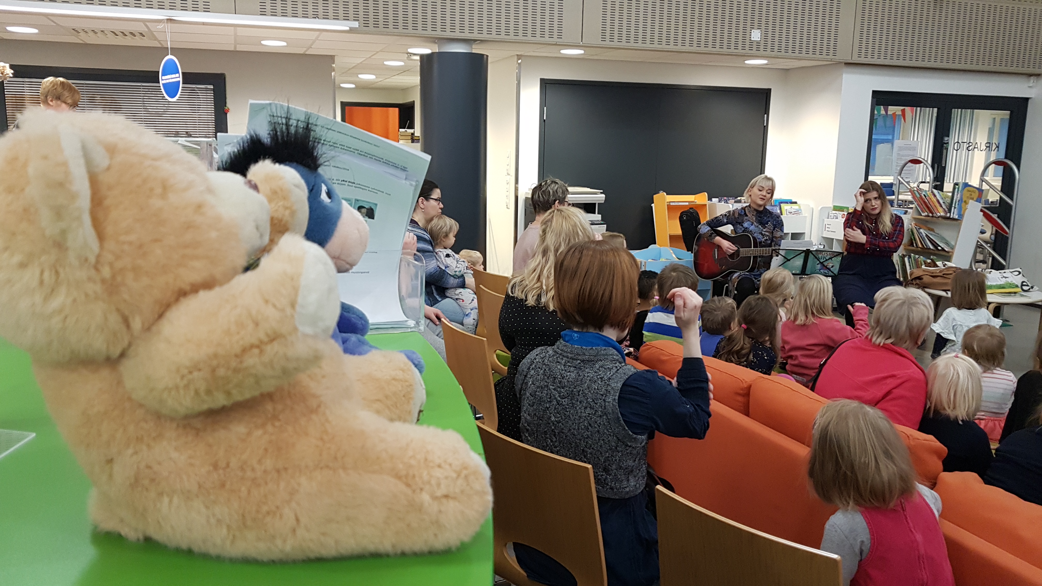 Kuvassa on musiikkitilaisuus kirjastossa. Esiintyjät soittavat kitaraa ja laulavat ja lapset osallistuvat tapahtumaan leikkien ja laulaen.
