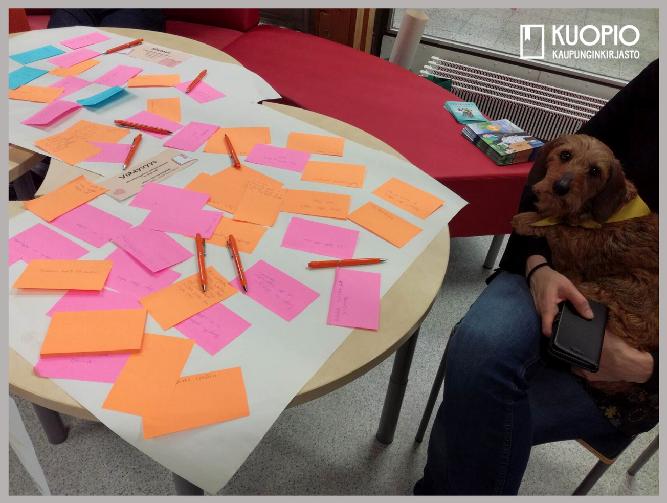Kuvassa on pöytä, jolla on paljon eri värisiä muistilappuja ja kyniä. Kuvassa on myös koira, joka lepää istuvan henkilön sylissä.